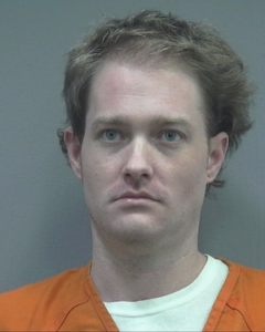 Mugshot of a man with light skin, short brown hair, and blue eyes. He is wearing an orange prison jumpsuit over a white shirt. His expression is neutral, and he is looking directly at the camera. The background is a plain, light-colored wall.