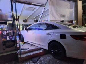 The image shows a white car that has crashed into a building, partially entering through what appears to be a glass storefront. The car is positioned with its front end inside the building, and there is visible damage to the glass and structure around it.