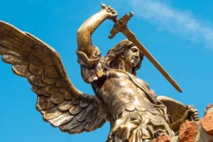 The image shows a statue of an angelic figure with large wings, holding a sword aloft in one hand. The statue is made of a material that resembles bronze or a similar metal, giving it a golden appearance. The angel has a determined expression and is wearing a flowing garment. The background is a clear blue sky with a few clouds.